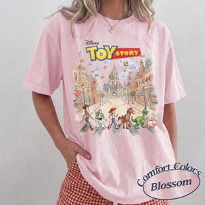 May include: A light pink Comfort Colors t-shirt featuring a Disney Toy Story graphic. The graphic shows the Toy Story characters walking down a street with a castle in the background. The shirt has short sleeves.