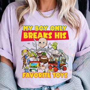 May include: A light purple t-shirt featuring a graphic print with the text "MY BOY ONLY BREAKS HIS FAVORITE TOYS" in yellow and red. The illustration depicts a collection of various disassembled and broken toy parts and action figures.