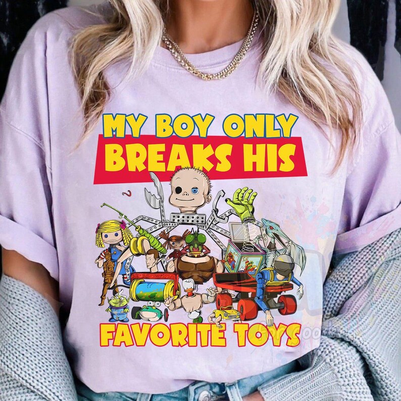 May include: A light purple t-shirt featuring a graphic print with the text "MY BOY ONLY BREAKS HIS FAVORITE TOYS" in yellow and red. The illustration depicts a collection of various disassembled and broken toy parts and action figures.