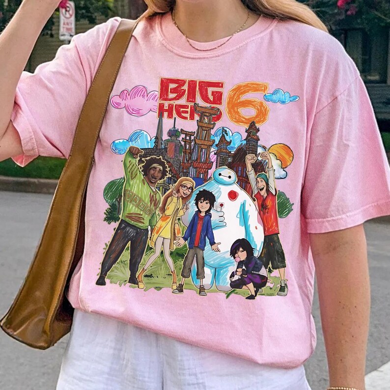 May include: A light pink t-shirt featuring a colorful graphic of characters and the text "BIG HERO 6". The design includes a cityscape, clouds, and the number 6. The shirt has short sleeves and a crew neck.