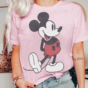 Peut inclure: Un t-shirt rose clair avec un grand dessin de Mickey Mouse. Mickey est repr&eacute;sent&eacute; dans sa pose classique, avec des oreilles noires, un visage blanc, un short rouge et des gants et chaussures blancs. Le t-shirt a des manches courtes et un col rond.
