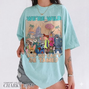 K&ouml;nnte beinhalten: Ein helles mintgr&uuml;nes T-Shirt mit kurzen &Auml;rmeln und einem Grafikdruck verschiedener animierter Tierfiguren aus Zootopia. Der Text "WE'RE WILD AND WE CAN'T BE TAMED" ist in einer fetten Retro-Schrift &uuml;ber und unter der Illustration gedruckt.