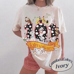 May include: Ivory-colored t-shirt featuring a graphic of the number "101" with Dalmatian dogs and characters from the animated film. The word "Dalmatians" is on a yellow banner. The shirt also has the words "Comfort Colors" and "Ivory".