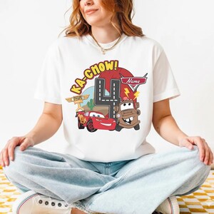 May include: White t-shirt featuring a colorful graphic of characters from the movie Cars, including Lightning McQueen and Mater. The design includes the number 4, the text "Ka-Chow!", and a space for a name. A fun birthday shirt.