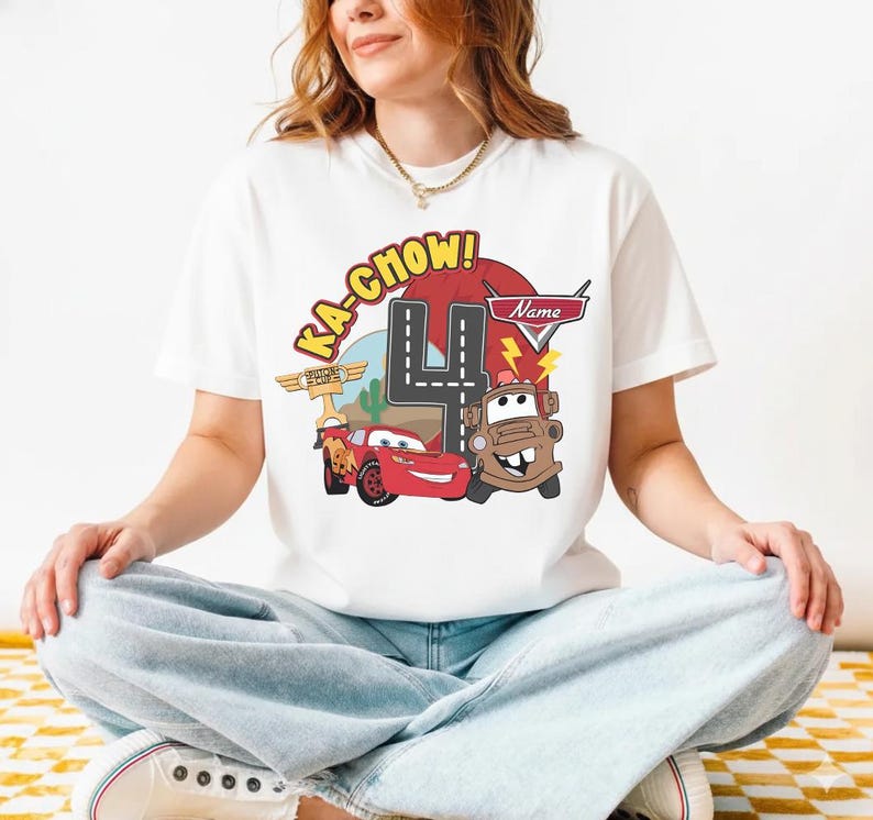 May include: White t-shirt featuring a colorful graphic of characters from the movie Cars, including Lightning McQueen and Mater. The design includes the number 4, the text "Ka-Chow!", and a space for a name. A fun birthday shirt.