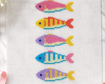 May include: A white needlepoint canvas featuring a vertical column of five stylized fish. Each fish is stitched in a different color combination, including pink, yellow, purple, and blue, with contrasting stripes and solid-colored tails.