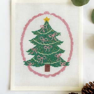 Puede incluir: Un diseño de árbol de Navidad bordado a punto de cruz. El árbol verde está decorado con lazos rosas y una estrella amarilla. El árbol está dentro de un marco ovalado rosa sobre un fondo blanco. El bordado está sobre un lienzo blanco.