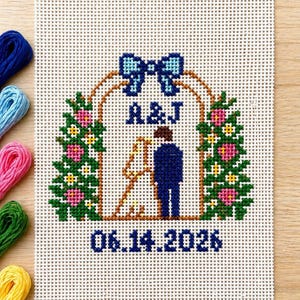 May include: Cross-stitch wedding design featuring a couple under an arch with the initials "A&J" and the date "06.14.2026". The design includes a blue bow, green foliage, and pink flowers. Colorful embroidery floss is visible on the left.