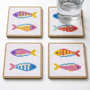 May include: Four square coasters with a cork border, each featuring a cross-stitched fish design. The fish are in various colors, including purple, yellow, blue, and red. A glass of water sits on one of the coasters.