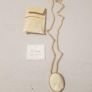 Peut inclure: Un collier pendentif camée crème avec un cadre doré, représentant le profil d'une femme. Le collier a un cordon torsadé et comprend une petite pochette et une étiquette Nina Ricci.