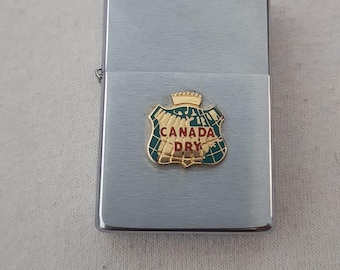 May include: A brushed silver metal lighter featuring a centered emblem with a gold crown above a green and gold grid shield. The text CANADA DRY is printed in red letters across the center of the shield, which is outlined in gold.