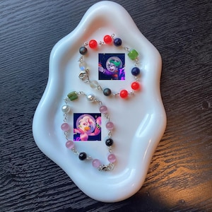 May include: Two beaded bracelets with colorful beads and small cartoon images are displayed on a white, wavy-edged dish. One bracelet features red, blue, green, and black beads. The other has pink, black, and pearl-like beads.