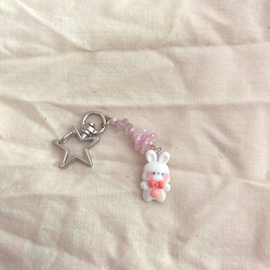 May include: A keychain featuring a white bunny charm with a pink bow and star-shaped beads. The keychain includes a silver star-shaped charm and a silver ring for attachment. The background is a light-colored fabric.
