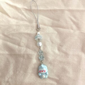 May include: A light blue phone charm with a silver loop. The charm features a string of beads, including a pearl, star-shaped beads, and a small blue figure with a red face and white belly.