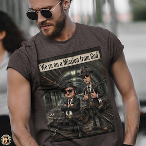 May include: Dark gray t-shirt featuring a graphic of two men in black suits and hats, with the text "We're on a Mission from God." The illustration depicts the men in a tunnel setting with a car.