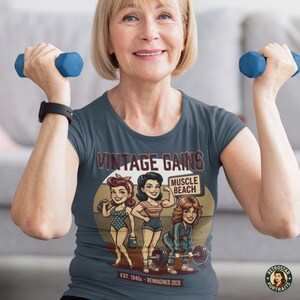 May include: A woman wearing a dark blue t-shirt with the words "VINTAGE GAINS" and a graphic of three women lifting weights. The shirt also has the words "MUSCLE BEACH" and "EST. 1940s - REIMAGINED 2026". The woman is holding two blue dumbbells.