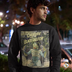 以下が含まれることがあります： 映画「Creature from the Black Lagoon」のヴィンテージスタイルのグラフィックが特徴の黒いスウェットシャツ。 グラフィックは、映画のタイトルがかすれたフォントで表示され、沼地環境にいるクリーチャーを描いています。 スウェットシャツは濃い色です。