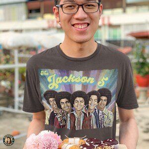 Może przedstawiać: Ciemnoszara koszulka z grafiką The Jackson 5, w stylu retro. Obraz przedstawia pięciu członków zespołu na kolorowym tle. Klasyczny t-shirt, idealny dla miłośników muzyki.