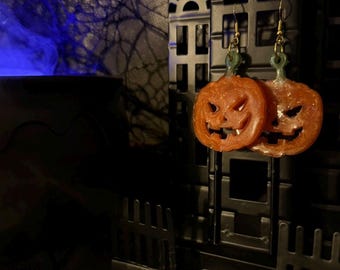 May include: A pair of orange pumpkin earrings with carved jack-o'-lantern faces. The earrings hang from gold-colored hooks and have green stems. The background includes a black decorative house and a dark, spooky setting.