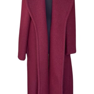 May include: A long, burgundy coat with a wide collar and a flowing silhouette. The coat is displayed on a black mannequin, showcasing its full length and design. The coat's color is a deep, rich shade of red.