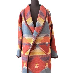 May include: A vibrant, open-front coat with a geometric pattern, reminiscent of Southwestern designs. The coat showcases a palette of red, orange, yellow, blue, and purple, with a brown trim. It is displayed on a black mannequin.