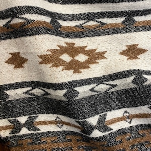 May include: Close-up of a woven textile with a geometric pattern. The design features alternating stripes of white, black, and brown. Brown diamond and arrow-like shapes are interspersed throughout the fabric, creating a textured look.