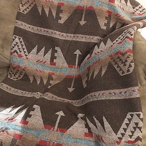 Op de afbeelding: Een bruine en beige wollen deken met een geometrisch patroon van pijlen en driehoeken. De deken heeft een geweven textuur en een gefranst randje.