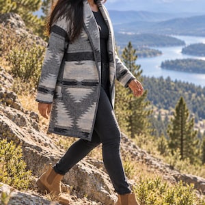 May include: A long-sleeved open-front coat featuring a gray, black, and white geometric pattern, worn over a black top and dark gray jeans, paired with tan suede ankle boots with elastic side panels and a low block heel.