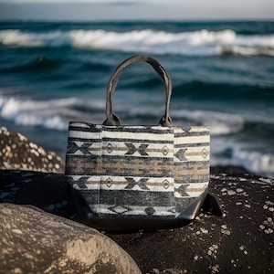 May include: A black and white patterned tote bag with brown accents. The bag has two black handles and a black bottom. The bag is sitting on a rocky surface.