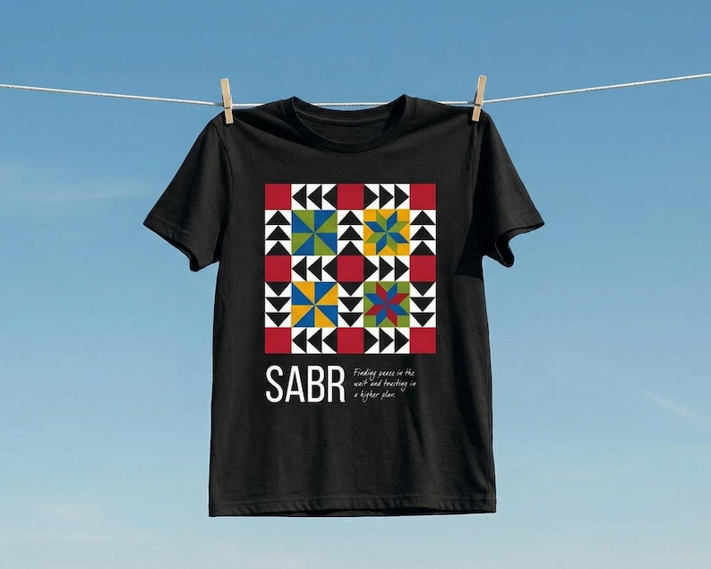 May include: Black t-shirt with a colorful geometric design. The design features red, yellow, blue, and green shapes arranged in a square pattern. The word "SABR" is printed in white, with the text "Finding peace in the wait and trusting in a higher plan."