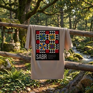 May include: A beige t-shirt with a colorful geometric design and the word "SABR" in white. The design includes red, blue, yellow, and black shapes. The text "Finding peace in the wait and trusting in a higher plan" is below the word "SABR".
