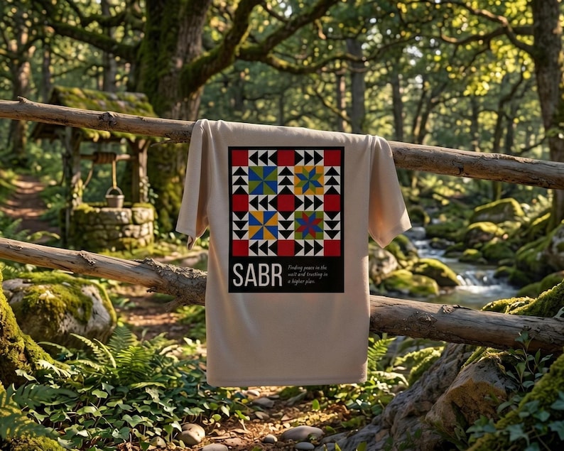 May include: A beige t-shirt with a colorful geometric design and the word "SABR" in white. The design includes red, blue, yellow, and black shapes. The text "Finding peace in the wait and trusting in a higher plan" is below the word "SABR".