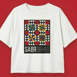 May include: White t-shirt featuring a colorful geometric design with red, blue, yellow, and green squares and triangles. The word "SABR" is printed in white on a black rectangle at the bottom of the design. Text below reads "trusting peace in the will and trusting in a higher plan."