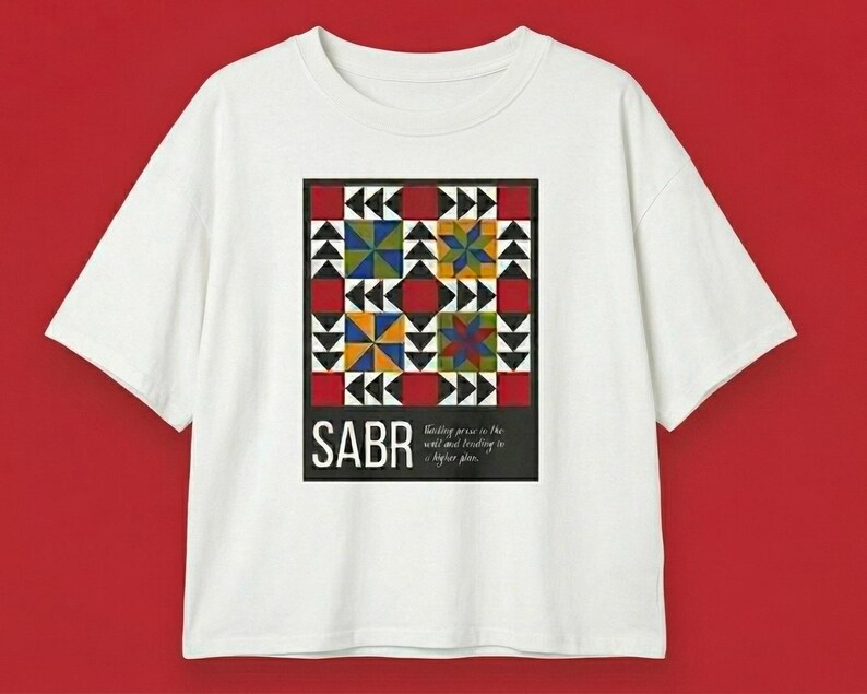 May include: White t-shirt featuring a colorful geometric design with red, blue, yellow, and green squares and triangles. The word "SABR" is printed in white on a black rectangle at the bottom of the design. Text below reads "trusting peace in the will and trusting in a higher plan."