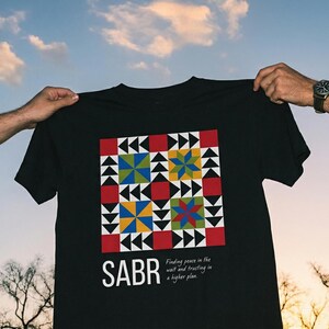 May include: Black t-shirt with a colorful geometric design and the word "SABR" printed on the front. The design includes red, yellow, blue, and green shapes. The text "Finding peace in the wait and trusting in a higher plan" is also printed on the shirt.