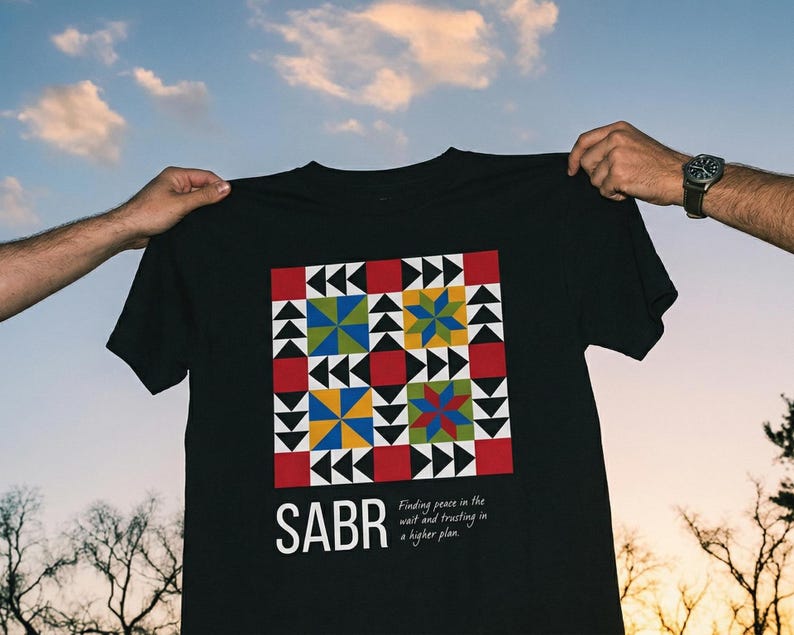 May include: Black t-shirt with a colorful geometric design and the word "SABR" printed on the front. The design includes red, yellow, blue, and green shapes. The text "Finding peace in the wait and trusting in a higher plan" is also printed on the shirt.