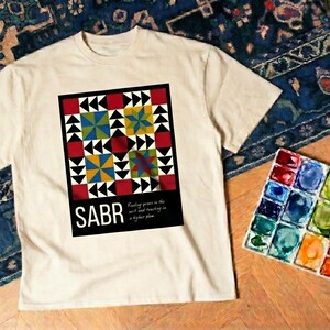 May include: Cream-colored t-shirt featuring a colorful geometric design with the word "SABR" in bold white letters. The design includes red, black, yellow, and blue shapes. A watercolor palette and brush are next to the shirt.