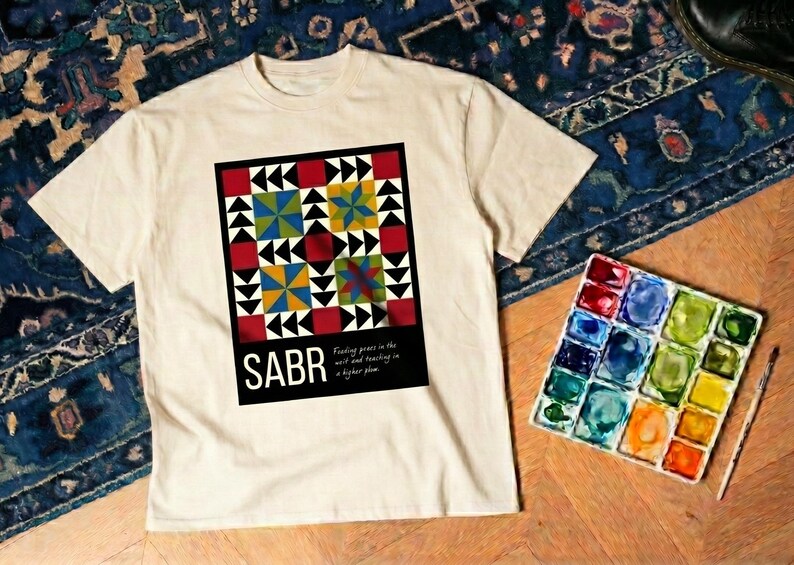 May include: Cream-colored t-shirt featuring a colorful geometric design with the word "SABR" in bold white letters. The design includes red, black, yellow, and blue shapes. A watercolor palette and brush are next to the shirt.