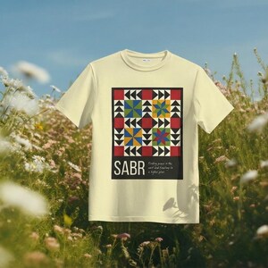 May include: A light yellow t-shirt featuring a colorful geometric design with red, black, blue, yellow, and green shapes. The word "SABR" is printed below the design, with the phrase "Finding peace in the self and trusting in a higher plan."