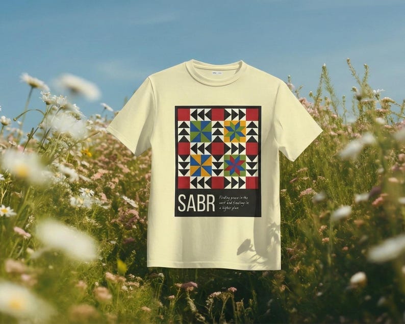 May include: A light yellow t-shirt featuring a colorful geometric design with red, black, blue, yellow, and green shapes. The word "SABR" is printed below the design, with the phrase "Finding peace in the self and trusting in a higher plan."