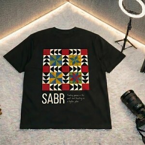 May include: Black t-shirt with a colorful geometric design and the word "SABR" in white. The design includes red, yellow, blue, and green shapes. The text "Finding peace in the wait and trusting in a higher plan" is also on the shirt.