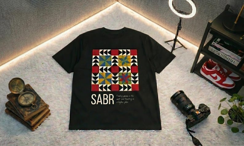 May include: Black t-shirt with a colorful geometric design and the word "SABR" in white. The design includes red, yellow, blue, and green shapes. The text "Finding peace in the wait and trusting in a higher plan" is also on the shirt.