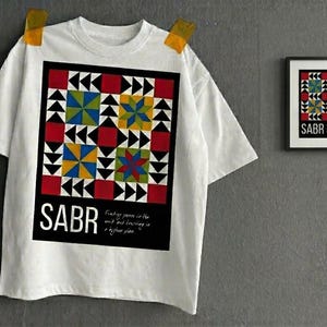 May include: White t-shirt with a colorful geometric design featuring red, blue, yellow, and green shapes. The word "SABR" is printed in large white letters at the bottom of the design. A framed print with the same design is on the right.