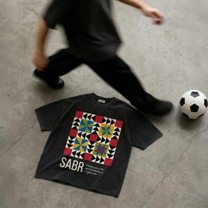 May include: A dark gray t-shirt with a colorful geometric design and the word "SABR" printed on the front. A soccer ball and a person in black clothing are also in the image.