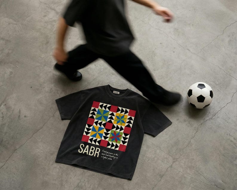 May include: A dark gray t-shirt with a colorful geometric design and the word "SABR" printed on the front. A soccer ball and a person in black clothing are also in the image.