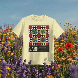May include: A light yellow t-shirt with a colorful geometric design and the word "SABR" in black. The design includes red, black, yellow, blue, and green shapes. The shirt is set against a backdrop of wildflowers and a blue sky.
