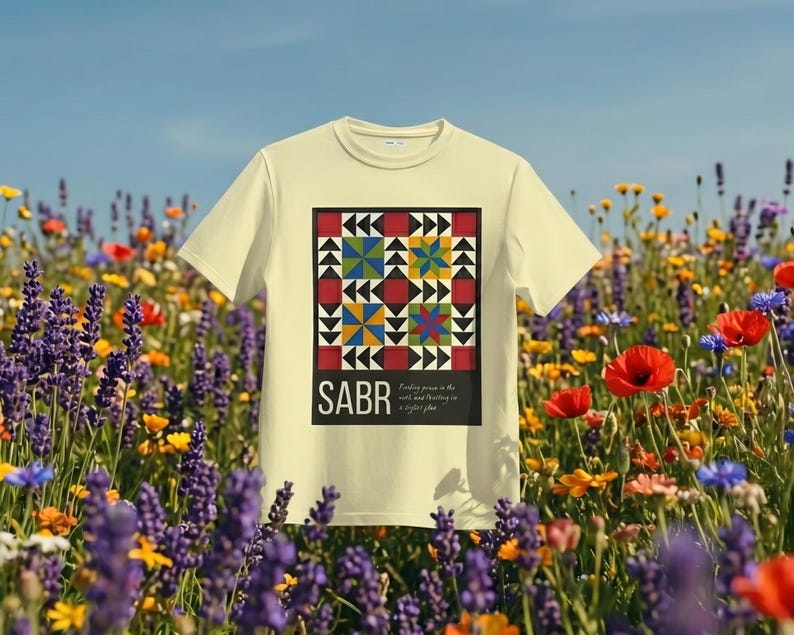 May include: A light yellow t-shirt with a colorful geometric design and the word "SABR" in black. The design includes red, black, yellow, blue, and green shapes. The shirt is set against a backdrop of wildflowers and a blue sky.