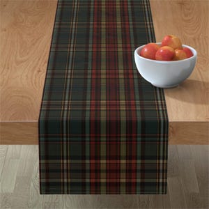 May include: A rectangular table runner featuring a dark green, red, and tan plaid pattern. A white bowl containing several red apples sits on the right side of the runner. The runner is laid out flat on a light brown wooden surface.