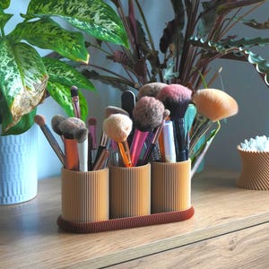 May include: A wooden makeup brush organiser with three sections, each holding different brushes. The light brown organiser has a ribbed design and sits on a dark brown base. Green and purple plants are in the background.