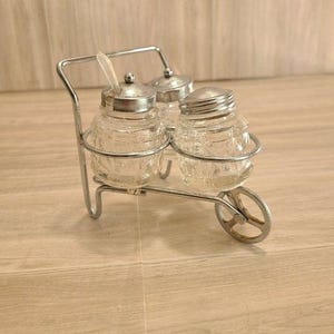 May include: A clear glass salt and pepper shaker set with a silver metal lid and a spoon, held in a decorative metal wheelbarrow-shaped holder. The glass jars have a textured pattern.